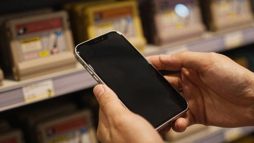 Hands inspecting a phone at the counter of a game shop, retro game cartridges on the shelves behind